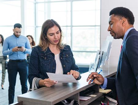 An adult male bank manager stands at the counter to answer the adult woman's questions about her monthly statements and recent check cashing.